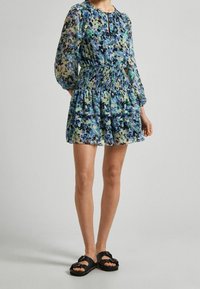 Kobieta w niebieskiej, kwiecistej sukience z długim rękawem, z marszczeniem w talii i falbanowym dołem, połączona z czarnymi sandałami na klamrę.