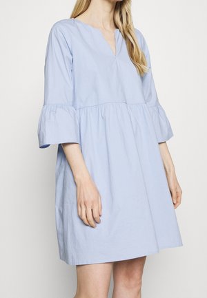 Nainen pukeutuneena vaaleansiniseen A-linjaisen helman mekkoon, jossa on kolmivartiset kellohihat ja V-muotoinen kaula-aukko, yksinkertaista vaaleaa taustaa vasten.