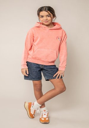 Fille debout, une jambe croisée, portant un sweat à capuche corail, un short en jean, des chaussettes blanches et des baskets orange et blanches sur un fond uni.