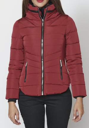 Frau trägt eine rote gesteppte Jacke mit Reißverschluss und seitlichen Reißverschlusstaschen sowie schwarze Hose, steht vor einem schlichten weißen Hintergrund.