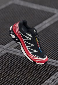 Schwarzer und roter Laufschuh mit strukturierter Oberfläche, ausgestattet mit einem GORE-TEX-Label, reflektierenden Akzenten und einem Schnellsystem für einen sicheren Sitz.