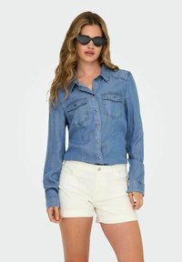 Chemise en denim avec boutons, deux poches poitrine, manches longues et couleur bleu clair, associée à un short blanc. Accessoirisée avec des lunettes de soleil noires.
