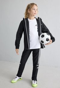 Veste de sport noire avec des accents blancs, associée à un pantalon noir avec des rayures blanches et une taille élastique. Tient un ballon de football.