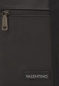 Zaino in nylon nero con tasca frontale con zip, finitura testurizzata e una patch in pelle con logo e testo argentato. Include spallacci resistenti.
