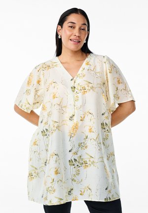 Tunique florale en tissu crème clair, avec un décolleté en V, des manches courtes et des motifs de fleurs jaunes et vertes sur l'ensemble.