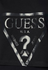Crna tkanina majica kratkih rukava s velikim metalik srebrnim trokutastim logotipom na kojem se nalaze tekstovi "GUESS", "U.S.A.", "WASHED" i dizajn upitnika.
