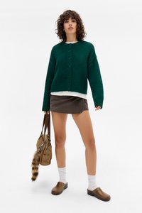 Cardigã tricotado verde com fecho de botão, usado sobre uma camisa branca, combinado com uma saia de veludo cotonço castanha e tamancos castanhos. Uma mala bege com um acento de pele.