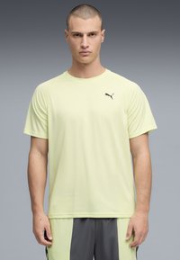 Άνδρας που φοράει ανοιχτό κίτρινο αθλητικό T-shirt Puma και δίχρωμο γκρι και κίτρινο σορτς, στέκεται μπροστά σε γκρίζο διαβαθμισμένο φόντο.