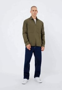 Olivgrüne Jacke mit zwei vorderen Taschen, mit Kragen, kombiniert mit dunkelblauen Jeans und weißen Sneakers, vor einem weißen Hintergrund.
