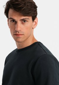 Svart sweatshirt med rund hals, som har en texturerad bomullsblandning, lätt figursydd design och synlig sömnad på axlarna.
