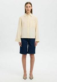 Camisa de botones amarilla claro con un bolsillo, combinada con pantalones cortos de mezclilla azul oscuro. La modelo lleva zapatos beige puntiagudos. Diseño simple y relajado.