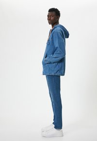 Chaqueta de denim con capucha, que presenta un bolsillo delantero y puños elásticos, llevada sobre pantalones de denim a juego y zapatos deportivos blancos.