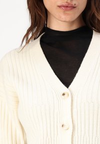 Cardigan a coste color crema con scollatura a V e due bottoni. Indossato sopra un top nero, con un motivo di maglia testurizzato.