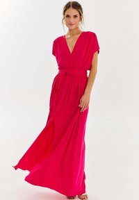 Długa różowa sukienka z lekkiego materiału. Posiada dekolt w kształcie litery V, krótkie rękawy oraz wiązanie w talii, z płynącą spódnicą i bocznym rozcięciem.