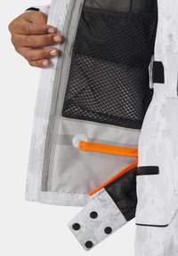 Intérieur de la veste isolée avec une poche en maille grise, un zip orange vif et un extérieur à motifs blancs. Présente un poignet à fermeture par pression.