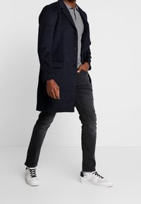 Navy wool coat with notched lapels, two buttons, and side pockets, paired with a gray polo, black jeans, and white sneakers.