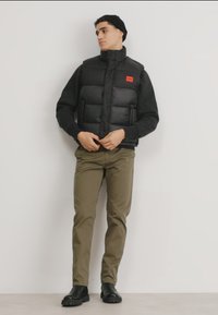Black padded vest with high collar, red logo patch, worn over a black long-sleeve shirt. Olive green pants and black boots complete the outfit.