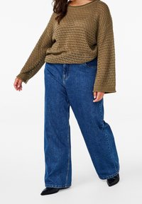 Khaki sweter robiony na drutach o luźnym splotem, z szerokimi rękawami, zestawiony z niebieskimi spodniami o szerokich nogawkach z wysokim stanem oraz czarnymi butami w szpic.