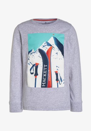 Chemise grise à manches longues avec un motif représentant des skis et des bâtons devant une montagne enneigée sous un ciel bleu avec un drapeau rouge.