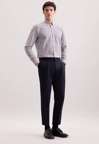 Camicia a quadri con bottoni in azzurro chiaro e bianco, abbinata a pantaloni scuri con taglio sartoriale e pieghe nette. Scarpe nere completano il look.