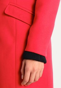 Abrigo rojo brillante con una textura suave, un corte entallado y un detalle negro de puño de punto acanalado que resalta el diseño de las mangas.