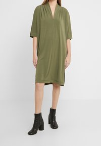 Olivgrön oversized klänning med V-ringning, korta ärmar och sidofickor, tillverkad av mjukt material. Kombinerad med svarta stövlar vid anklarna.