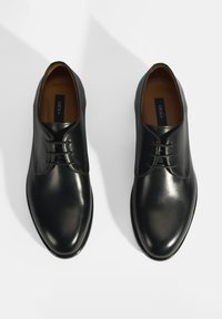 Chaussures en cuir noir avec une finition lisse, bout rond, design à lacets et détails de couture subtils sur le dessus. La semelle est plate.