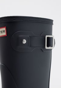 Botte de pluie en caoutchouc noir avec accent de boucle, haut arrondi et tige texturée. Détail du logo sur une étiquette rouge près du haut.