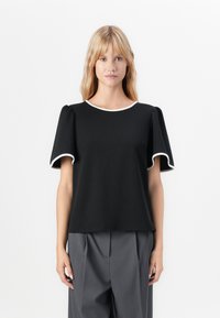 Femme portant un haut noir à manches courtes avec des bordures blanches et un pantalon plissé gris, se tenant contre un fond blanc uni.