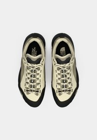 Világos bézs sportcipők fekete részletekkel, texturált felsőrész, rugalmas gumitalp és logó a talpbetéten; mintás cipőfűzőkkel és megerősített orral.