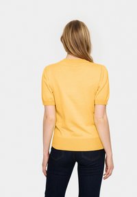Žlutá blůza s krátkým rukávem a kulatým výstřihem, se zamotanými rukávy a přiléhavým pasem, vyrobená z měkkého pleteného materiálu.