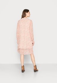 Vestido floral fluido em rosa claro e branco, com mangas longas, punhos com babados e design em camadas. Usado com botas de tornozelo castanhas.