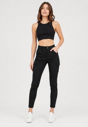 Donna con top corto nero senza maniche, pantaloni cargo neri, sneakers bianche, in piedi con una mano in tasca su sfondo semplice.