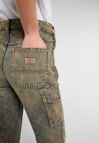 I pantaloni cargo in denim marrone chiaro presentano una grande tasca posteriore con un'etichetta cucita. Un leggero effetto invecchiato aggiunge texture al tessuto.
