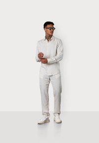 Eine Person steht vor einem schlichten Hintergrund, trägt ein gestreiftes passendes Outfit und eine Sonnenbrille, hat eine selbstbewusste Körperhaltung und einen entspannten Ausdruck.