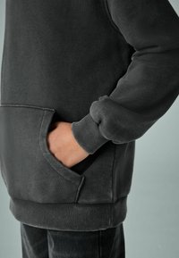 Tamsiai pilkas džemperis su kengūros kišene, nėriniais rankovėse ir apačioje, pagamintas iš minkštos medžiagos su lygiu tekstūru.