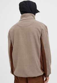 Brun og beige fleecejakke med teksturert stoff, høy krage og avslappet passform. Har kontrasterende brune sidepaneler.