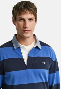 Blauw en marine gestreept rugbyshirt met een grijze kraag, voorzien van drie knopen en een klein logo op de borst. Van katoen.