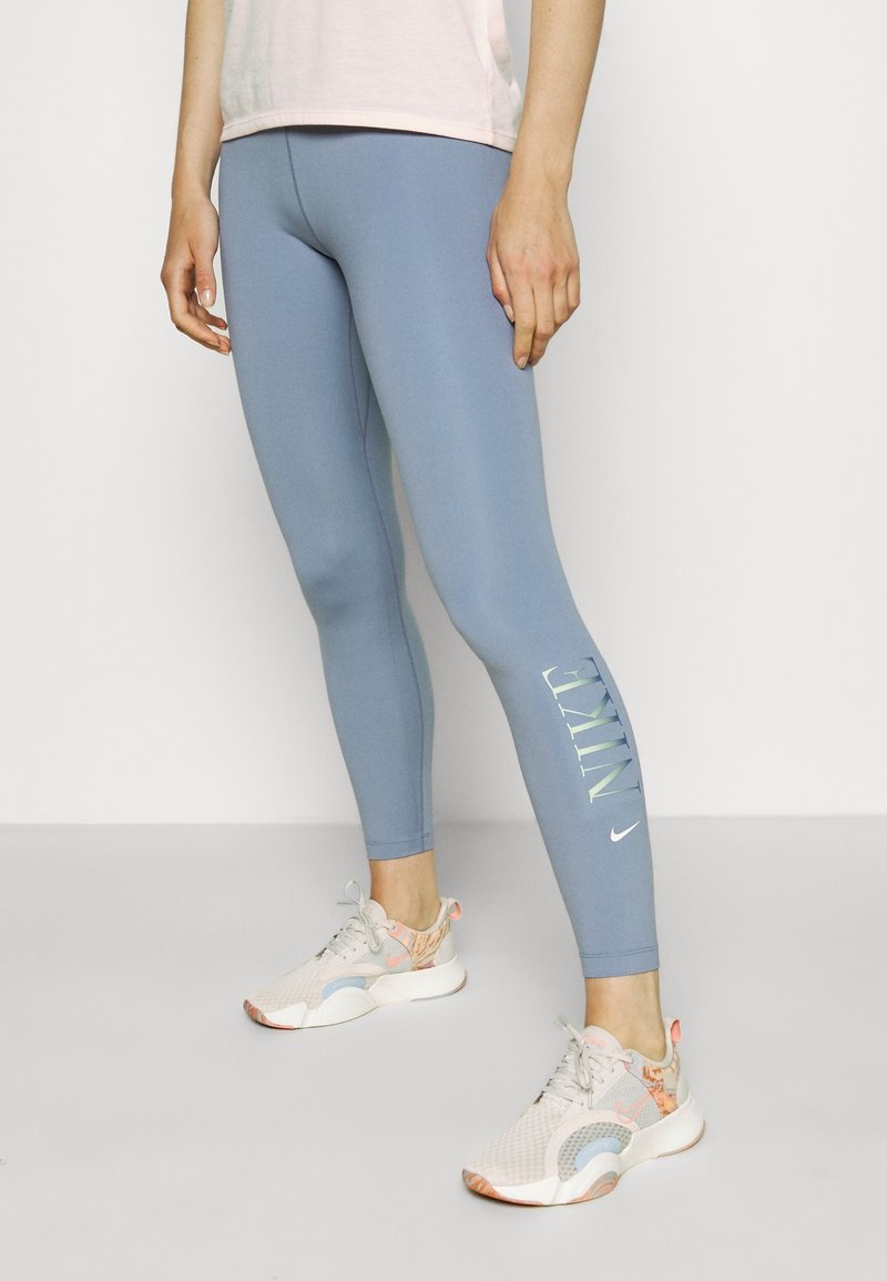 Leggings ceñidos de color azul con el logotipo "NIKE" impreso en verde claro. Llevados con zapatillas claras, con una camiseta rosa visible arriba.
