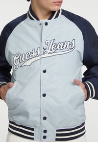 Lichtblauwe nylon bomberjack met marineblauwe mouwen en geribbelde gestreepte manchetten. Bevat het "Guess Jeans"-logo in witte borduursel en drukknoopsluitingen.