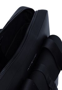 Borsa in tessuto nero con chiusura a zip, caratterizzata da una texture liscia e una tracolla laterale. Design minimalista con dettaglio del logo sul lato.