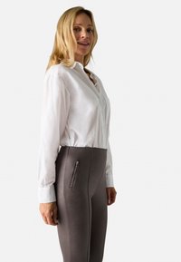 Camisa blanca de botones con mangas largas combinada con pantalones grises de tiro alto y stretch que cuentan con un bolsillo con cremallera lateral y una textura suave.