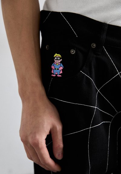 Pantaloni din denim negri cu cusături geometrice albe; prezintă un patch brodat colorat cu un personaj pe buzunarul din față.