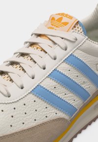 Λευκό αθλητικό παπούτσι Adidas με ανοιχτό μπλε ρίγες, διάτρητο δέρμα, μπεζ δερμάτινη μύτη και πορτοκαλί λογότυπο στη γλώσσα.