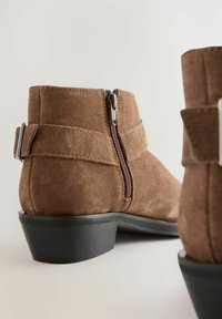 Botas de tobillo de ante marrón con una cremallera lateral y una correa decorativa, con un tacón negro apilado y una textura suave.