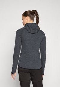 Giacca sportiva grigia con maniche lunghe e cappuccio. Presenta una vestibilità slim, una texture liscia e cuciture a contrasto. Indossata con pantaloni scuri.