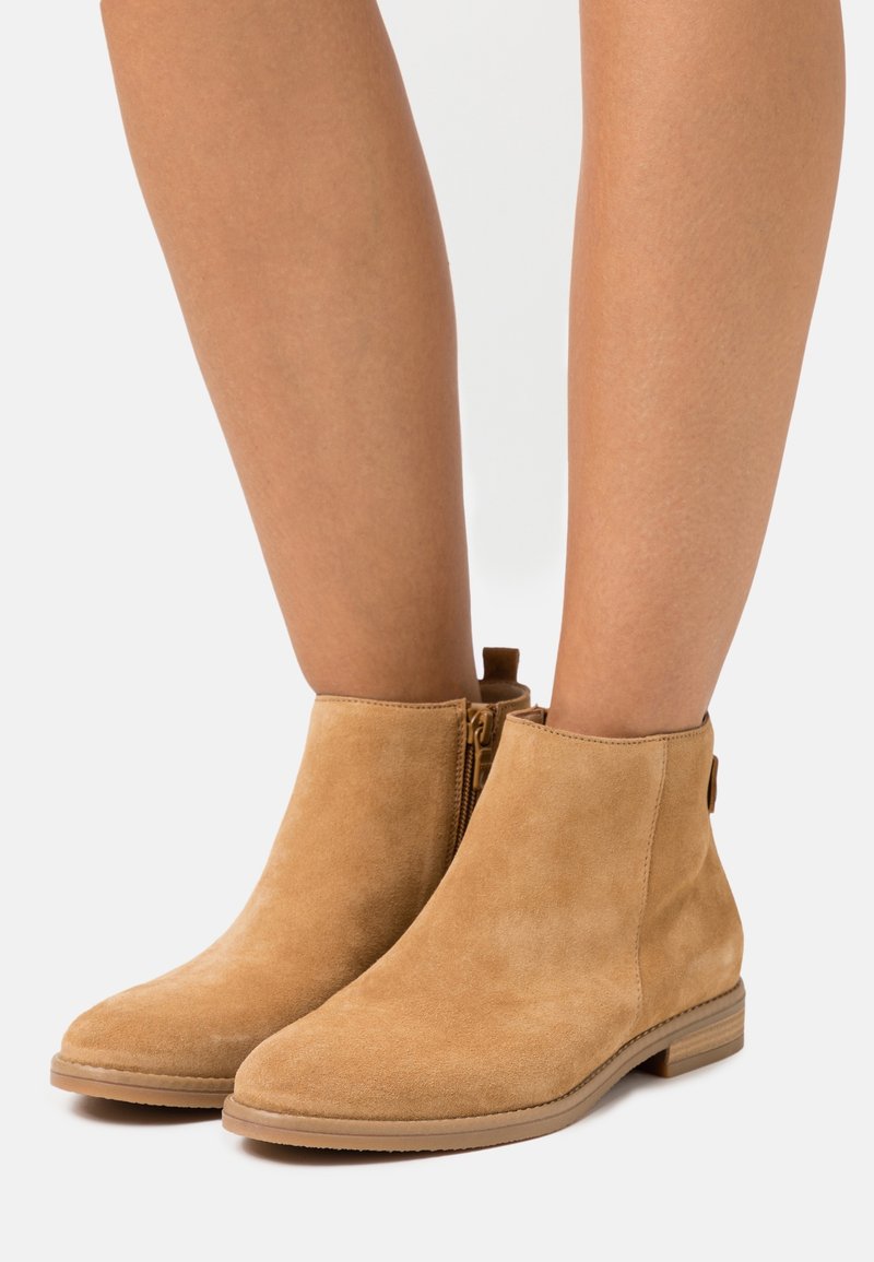 Bottines en daim marron clair avec fermeture éclair latérale, portées par une personne aux jambes nues, sur fond blanc.