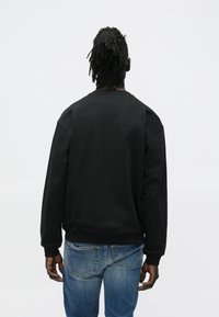 Μαύρο φλις φούτερ με στρογγυλή λαιμόκοψη, χαλαρή εφαρμογή και ριμπ μανσέτες. Φορώντας το με μπλε τζιν, θέα από πίσω.