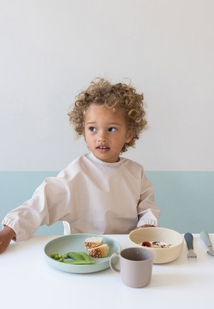 Copil tânăr cu păr creț, așezat la o masă cu o farfurie cu pâine și mazăre, un bol cu cereale cu fructe de pădure și o ceașcă.