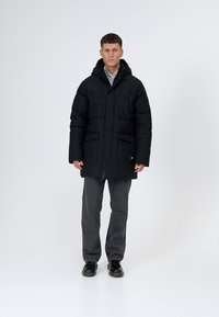 Black puffer jacket with a hood, featuring a zip front, two chest pockets, and two lower pockets, paired with gray pants and black shoes.